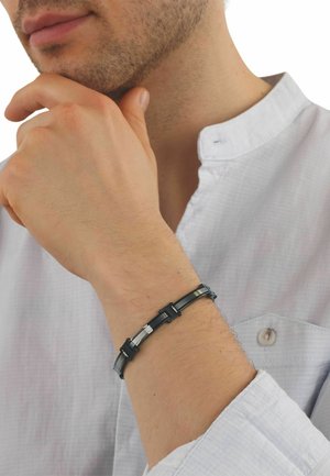 Uomo con camicia bianca che indossa un braccialetto metallico nero e argento, mano appoggiata pensierosa vicino al mento.