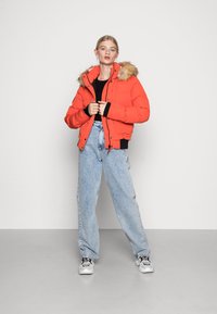 Oranje pufferjack met een met bont gevoerde capuchon, voorzien van een rits aan de voorkant, geribde manchetten en een cropped ontwerp, gecombineerd met losse lichtblauwe denim jeans.