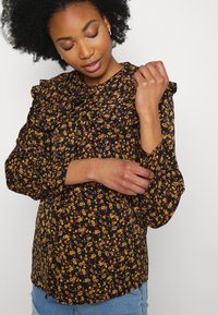 Blusa preta com padrão floral, com gola e mangas em renda, apresentando flores amarelas e laranjas, e um tecido texturizado. Combinada com denim.