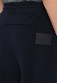 Navyblauwe sweatpants van textuurstof, met een elastische tailleband en een zwart rubberen logo met de tekst 'EA7 Emporio Armani' op de rug.