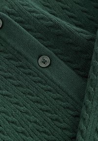 Tmavo zelený pletený kardigán s textúrovaným vzorom z kábla, okrúhly čierny gombík s logom "Clarks" a hladkými rebrovanými manžetami.