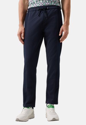 Man wearing navy drawstring pants, white sneakers with green laces, and a patterned shirt, standing with hands relaxed by sides.