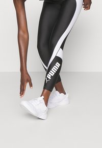 Black athletic leggings with white side stripes, featuring a large "PUMA" logo on the calf, paired with white sports shoes.