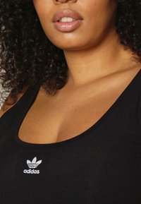 Top in maglia nera con coste, caratterizzato da un logo Adidas ricamato in bianco sul petto. Texture liscia con scollo a girocollo e spalline larghe.