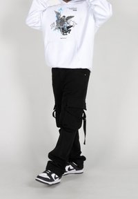 Sudadera blanca con estampado gráfico, pantalones cargo negros con bolsillos laterales y correas, y zapatillas en blanco y negro. Fondo liso.