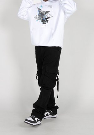 Felpa bianca con stampa grafica, pantaloni cargo neri con tasche laterali e cinghie, e sneakers bianco e nere. Sfondo semplice.