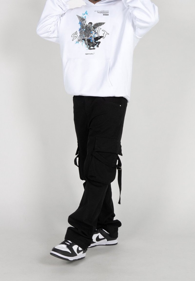 Sudadera blanca con estampado gráfico, pantalones cargo negros con bolsillos laterales y correas, y zapatillas en blanco y negro. Fondo liso.