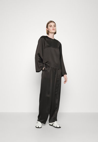 Haut noir en satin à manches longues avec un col rond, associé à un pantalon noir à jambes larges. Le mannequin porte des sandales blanches avec un design entrecroisé.