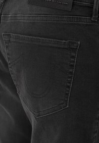 Lähikuva mustista denimpükstest, mille tagakotis on tikitud hobuserauakujuline disain ja naha silt "True Religion" kotikese kohal.