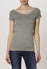 Šedé tričko s krátkým rukávem a širokým výstřihem, zdobené prošívaným lemem. Hladká textura a slim fit, v kombinaci s modrými džínami.