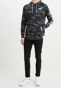 Svart camouflage-hoodie med vita dragkedjor och logga, kombinerad med svarta tapered-byxor och vita sneakers. Slät tygkvalitet och avslappnad passform.