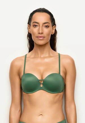 Donna con capelli lunghi e scuri che indossa un reggiseno verde senza spalline con sottili spalline, rivolta in avanti su uno sfondo semplice.
