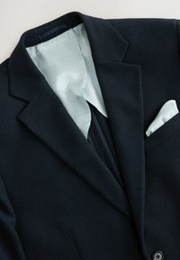 Granatowa marynarka o teksturowanej powierzchni z wzorzystą jasnoniebieską podszewką, klapami w stylu notch oraz białą chusteczką w kieszeni. Posiada dwa czarne guziki oraz dopasowany krój.