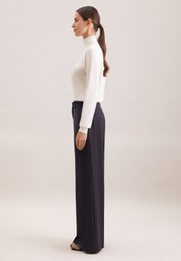 Biała bluzka z długim rękawem i wysokim kołnierzem połączona z czarnymi, dopasowanymi spodniami o szerokich nogawkach. Stylizacja charakteryzuje się gładką teksturą i dopasowanym fasonem.
