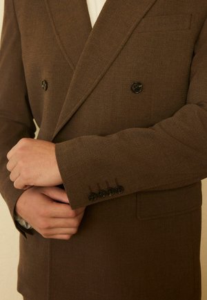 Mann in braunem zweireihigem Blazer passt die Manschette an und zeigt dunkle Knöpfe sowie strukturierten Stoff vor neutralem Hintergrund.