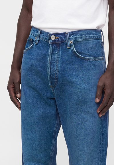 Gros plan d'une personne portant un jean bleu taille haute avec une fermeture par bouton et des poches avant, associé à une chemise blanche.