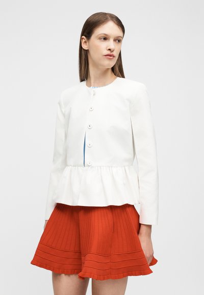 Γυναίκα που φοράει λευκό σακάκι με κουμπιά και πεπλούμ και πορτοκαλί πλισέ φούστα μήκους μέχρι το γόνατο, κοιτάζει πλάγια σε απλό φόντο.