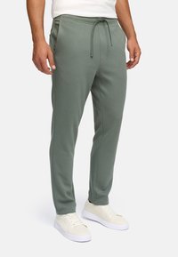 Mand iført sagegrønne joggers med snørebånd og hvide sneakers, stående foran en ensfarvet hvid baggrund.