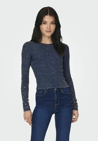 Maglietta a maniche lunghe blu navy con texture a coste, dettagli in perle sulle maniche e un design aderente, abbinata a jeans scuri a vita alta.