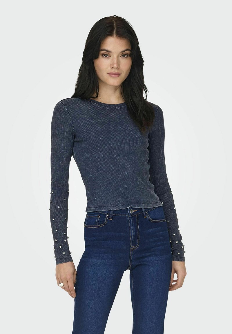 Maglietta a maniche lunghe blu navy con texture a coste, dettagli in perle sulle maniche e un design aderente, abbinata a jeans scuri a vita alta.