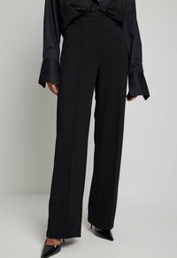 Schwarze hochtaillierte Hose mit geradem Schnitt, vorne gefaltet, aus glattem Material und mit einem Bund. Kombiniert mit spitzen schwarzen Heels.