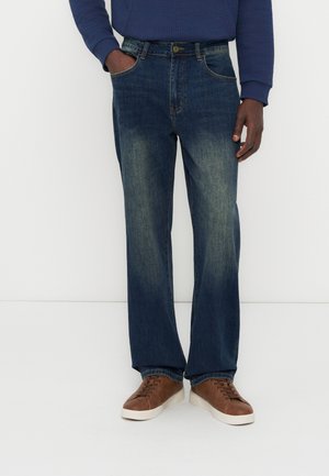 Άνδρας που φοράει ξεθωριασμένα μπλε τζιν με ίσια γραμμή, μπλε μπλούζα με μακριά μανίκια και καφέ αθλητικά παπούτσια με λευκές σόλες, στέκεται με το ένα χέρι στην τσέπη.