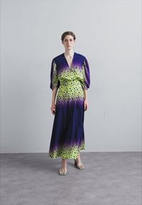 Maxi jurk met een diepe V-hals, pofmouwen, verloop van paars naar groen met zwarte vlekken, gecombineerd met groene sandalen.