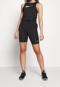 Czarny sportowy top na ramiączkach Nike z logo i detalem w postaci węzła, zestawiony z czarnymi szortami rowerowymi i czarnymi butami sportowymi. Gładka tekstura, dopasowany krój.