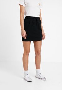 Gonna nera con vita alta, cordoncino regolabile e design plissettato. Indossata con una maglietta bianca e sneaker bianche per un look casual.