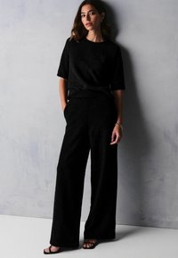 Černý strukturovaný dvoudílný outfit s krátkým rukávem a širokými kalhotami, stylizovaný s náramkem a sandály na řemínky.