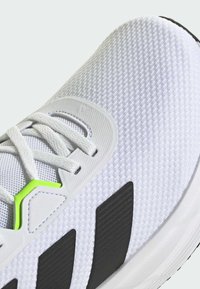 Zapatilla deportiva blanca con parte superior de malla texturizada, franjas laterales negras y un acento verde cerca de los cordones. Presenta una tapa de talón moldeada.
