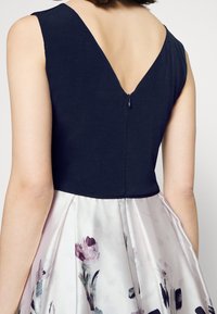 Námořnický top s designem ve tvaru V na zádech, doplněný květinovou plizovanou sukní ve světle růžových a levandulových odejích, s hladkým saténovým povrchem.