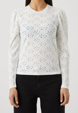 Vrouw draagt een witte top met lange mouwen met oogjesbloemenpatroon en opgepofte schouders, gecombineerd met een zwarte broek.