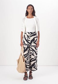 Cardigan en crochet blanc sur un top blanc, associé à un pantalon large à rayures zèbres noir et crème. Tenant un sac fourre-tout tissé beige.