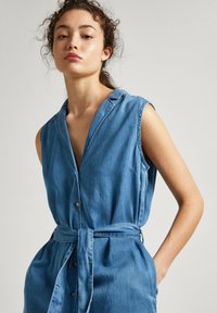 Robe sans manches en denim avec un décolleté en V, des boutons à l'avant et une taille à nouer. Elle possède un tissu bleu clair doux et des poches latérales.