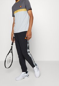 Osoba v šedém a žlutém sportovním tričku, černých teplácích se pruhy, bílých teniskách Lacoste, drží černou tenisovou raketu.