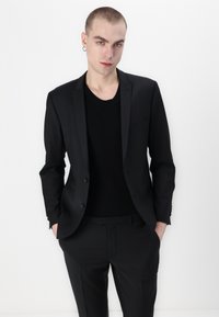 Zwarte op maat gemaakte colbert over een zwart T-shirt met ronde hals, met slim-fit broek. Glad materiaal, minimalistisch ontwerp, sluiting met twee knopen.