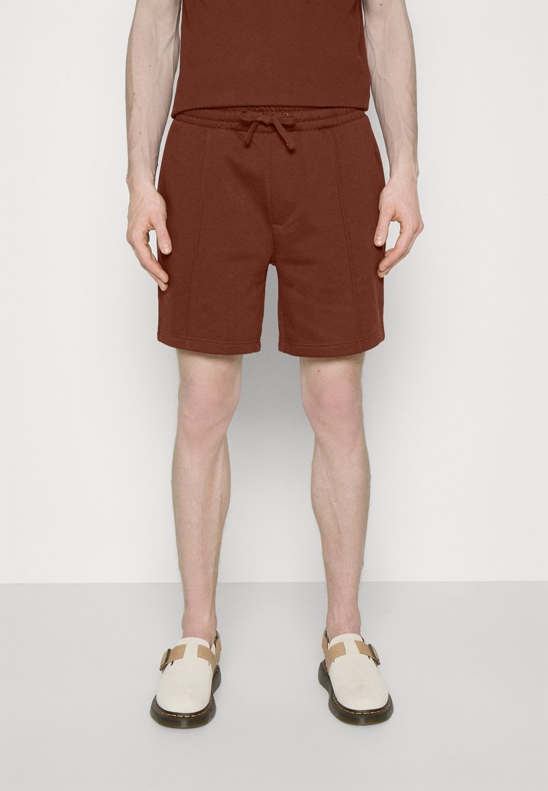 Homme portant un short marron à cordon et un top sans manches assorti avec des chaussures beige à enfiler sur un fond uni.