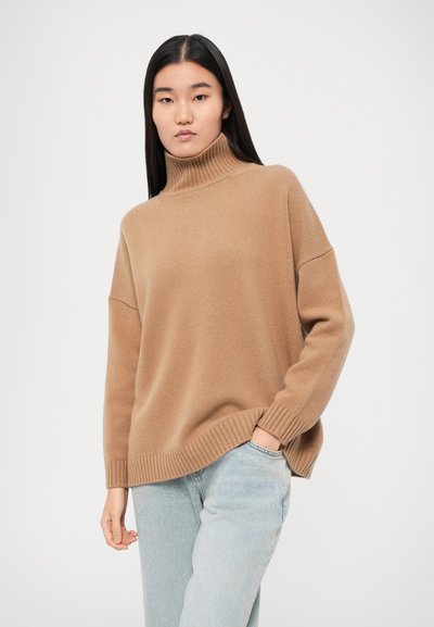 Pull encolure roulée beige avec texture côtelée, épaules tombantes et manches à revers, associé à un jean bleu clair. Design simple et décontracté.