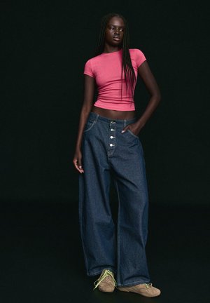 Ženska z dolgimi pletenimi lasmi, oblečena v oprijet vrh rožnate barve, široke kavbojke z visokim pasom in bež superge z rumenimi vezalkami.