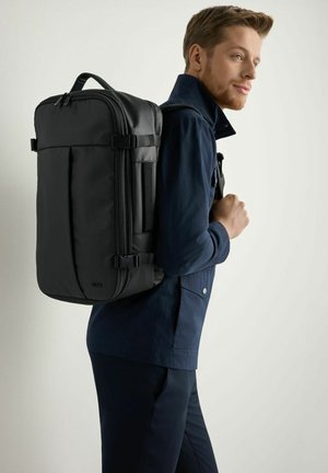 Mężczyzna w granatowym stroju niosący duży czarny prostokątny plecak, stojący bokiem na jednolitym jasnym tle.