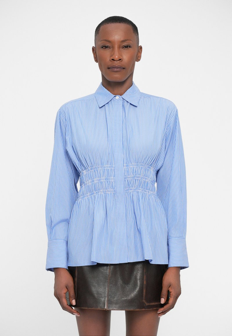 Camicia a righe azzurro chiaro con vita aderente, maniche lunghe e orlo a peplum. Presenta un collo classico e dettagli arricciati in vita.