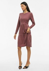 Långärmad mauve klänning med figurnära bodice och rynkad, flerlagers kjol. Slät tygtextur med rund halsringning och längd nedanför knäna.