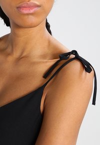 Svart klänning med tunna band och rosetter, mjuk textur och en figurnära design som framhäver halslinjen och axelområdet.