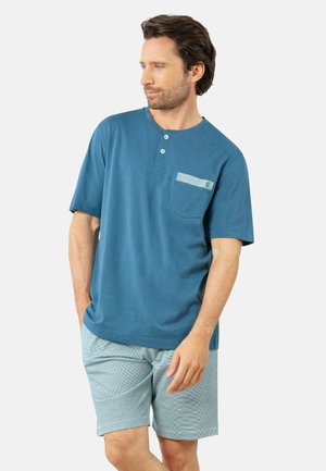 Homme en chemise henley bleu à manches courtes avec poche poitrine et short bleu clair à motifs, debout les mains dans les poches, regardant vers la gauche.
