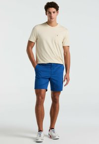 Lichtbeige t-shirt met korte mouwen en een borstzak, gecombineerd met blauwe shorts; het model draagt witte sneakers met rode en blauwe accenten.
