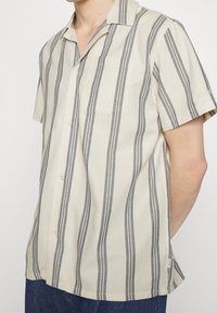 Camisa de manga corta con botones en beige con rayas verticales grises, que cuenta con un bolsillo en el pecho y un diseño de cuello relajado.