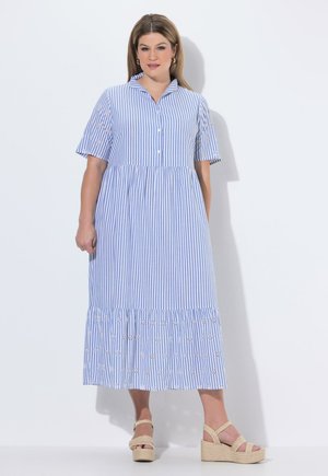 Kobieta stojąca, ubrana w niebiesko-białą sukienkę midi w paski z haftowanymi kwiatowymi detalami oraz beżowe sandały na koturnie na jednolitym białym tle.