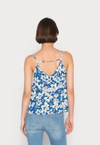 Blå blommig ärmklänning med vita blommönster, justerbara spaghetti-stroppar och rundad ryggdesign. Matchad med ljusblå jeans.