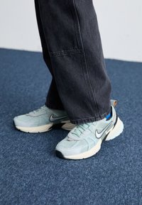 Zapatillas Nike de malla azul claro con un acabado texturizado, swoosh negro y suela media crema, combinadas con pantalones de pierna ancha gris oscuro.